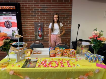 Emma Copley standing in front of Alpha Omicron Pi's homecoming booth in the university center. The table is bright yellow with touches of pink and white.